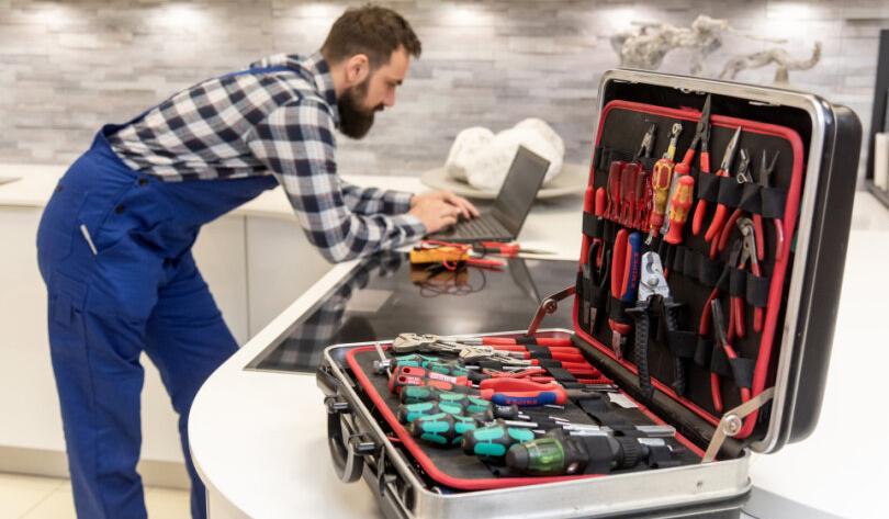 Handwerker steht in Küche und arbeitet am Laptop, Werkzeugkoffer mit Werkzeugen geöffnet.