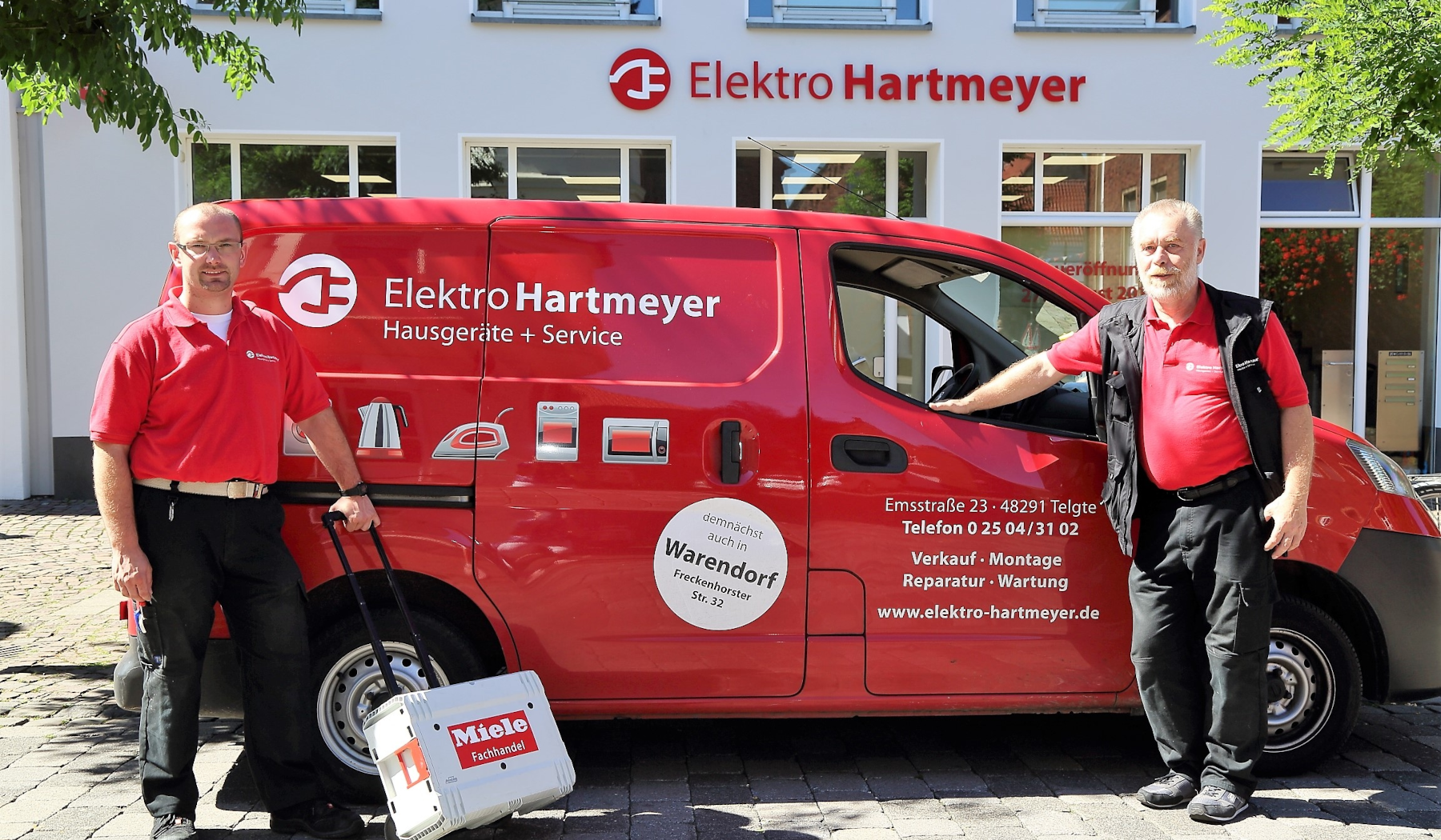 Zwei Männer in roten Arbeitshemden stehen vor einem roten Elektrofirmenwagen.